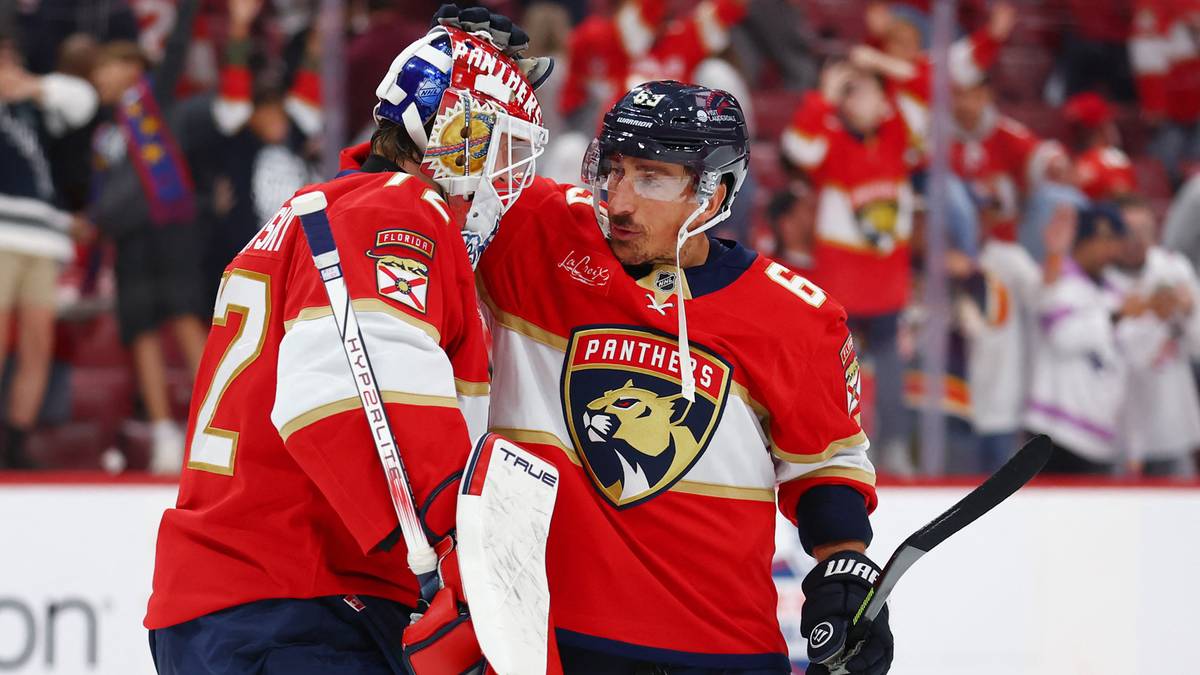 Dwóch hokeistów w czerwonych strojach, jeden w kasku bramkarskim, rozmawia ze sobą na lodowisku.