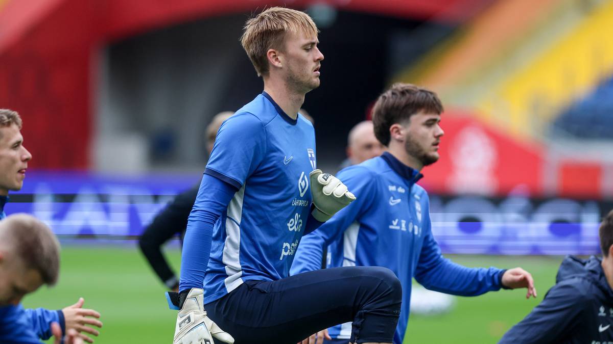 Dwóch piłkarzy w niebieskich strojach treningowych, jeden z nich bramkarz, wykonuje rozgrzewkę na murawie stadionu.