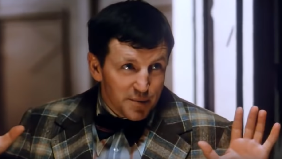 Portret mężczyzny w garniturze w kratę, z lekko pochyloną głową i uniesionymi dłońmi. Mężczyzna ma ciemne włosy i patrzący w bok wzrok.