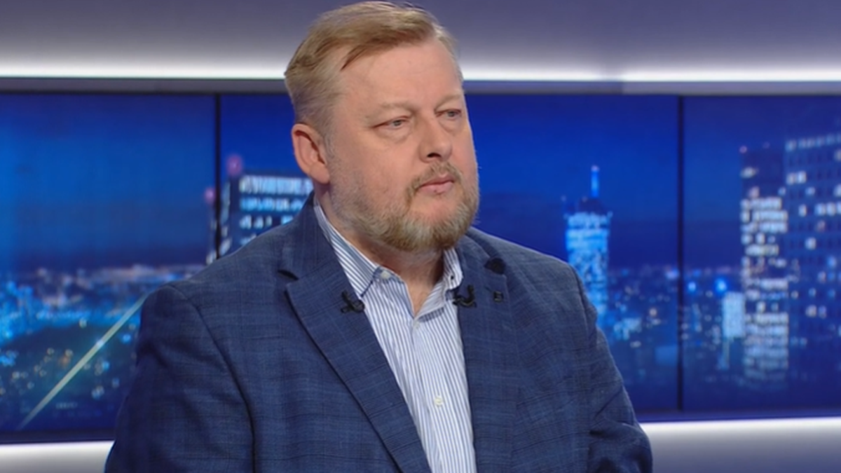 Mężczyzna w niebieskiej marynarce siedzi przed ekranem z nocnym widokiem miasta.