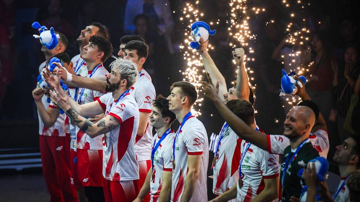 Grupa siatkarzy w strojach sportowych z medalami na szyjach, trzymających w rękach niebieskie czapki z białymi pomponami. W tle widać fajerwerki i tłum widzów.