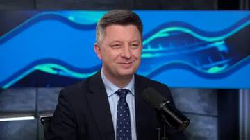 Mężczyzna w garniturze siedzi przed mikrofonem, uśmiechając się.