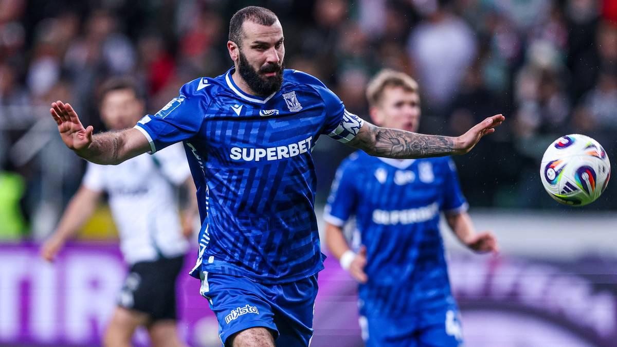 Zawodnik Lecha Poznań w niebieskiej koszulce z logiem "SUPERBET" rozkłada ręce na boki, gotowy do akcji.