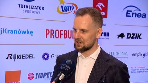 Mężczyzna z zarostem i w ciemnym garniturze, z mikrofonem przed ustami, stoi na tle ścianki z wieloma logotypami sponsor&oacute;w, m.in. LOTTO, Enea, Riedel.