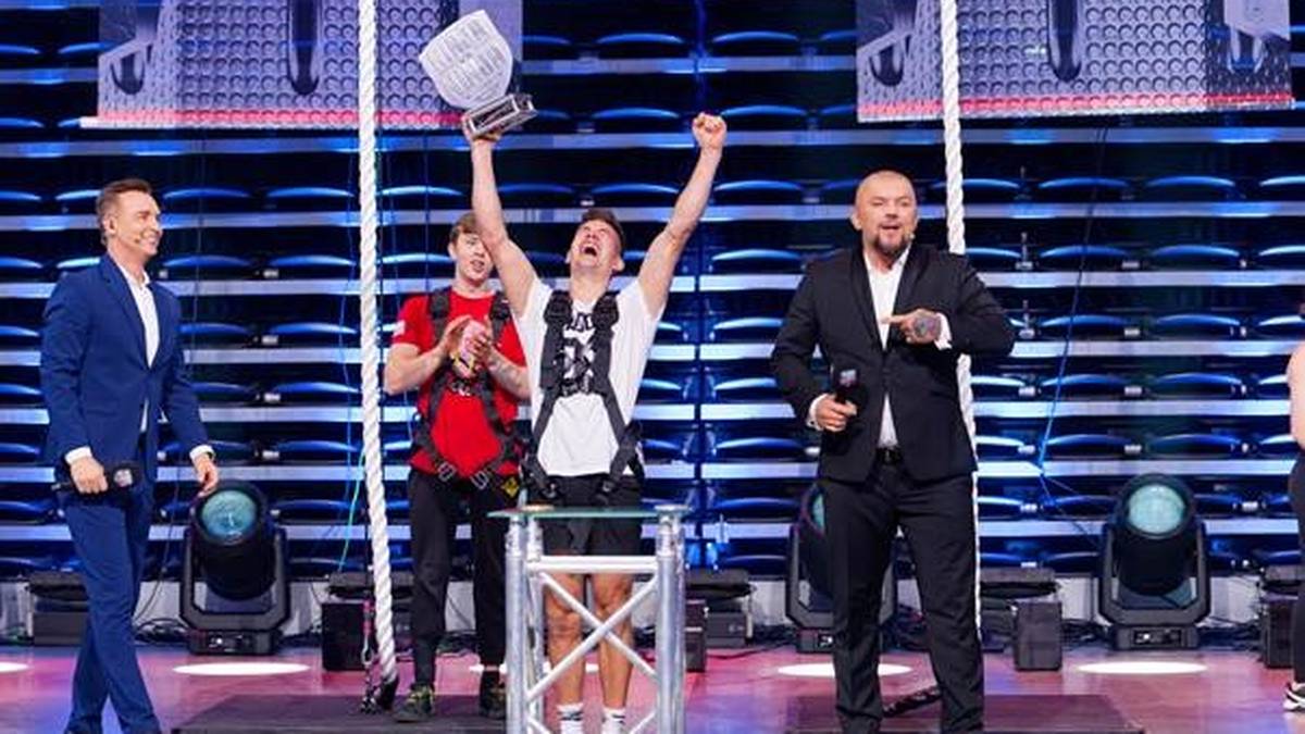 Szczęśliwy mężczyzna w białej koszulce z trofeum w uniesionej dłoni, podczas finału zawod&oacute;w.