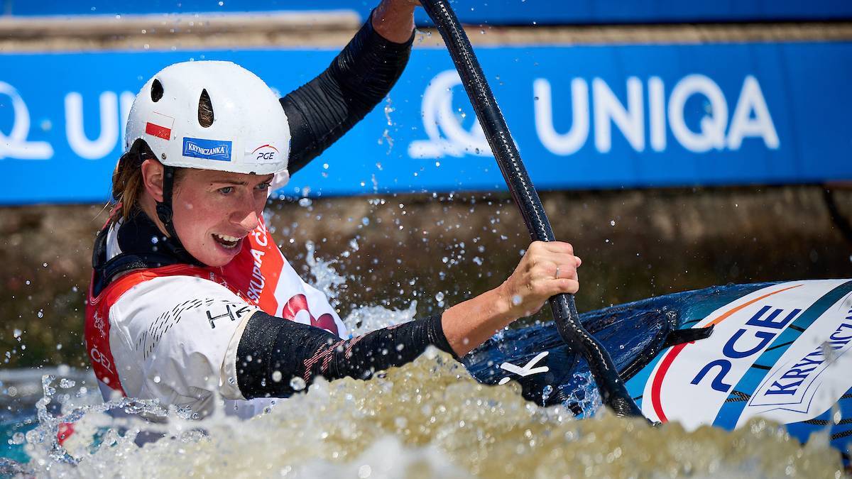 Kobieta w kasku i kamizelce sportowej wiosłuje w kajaku, ochlapując się wodą.