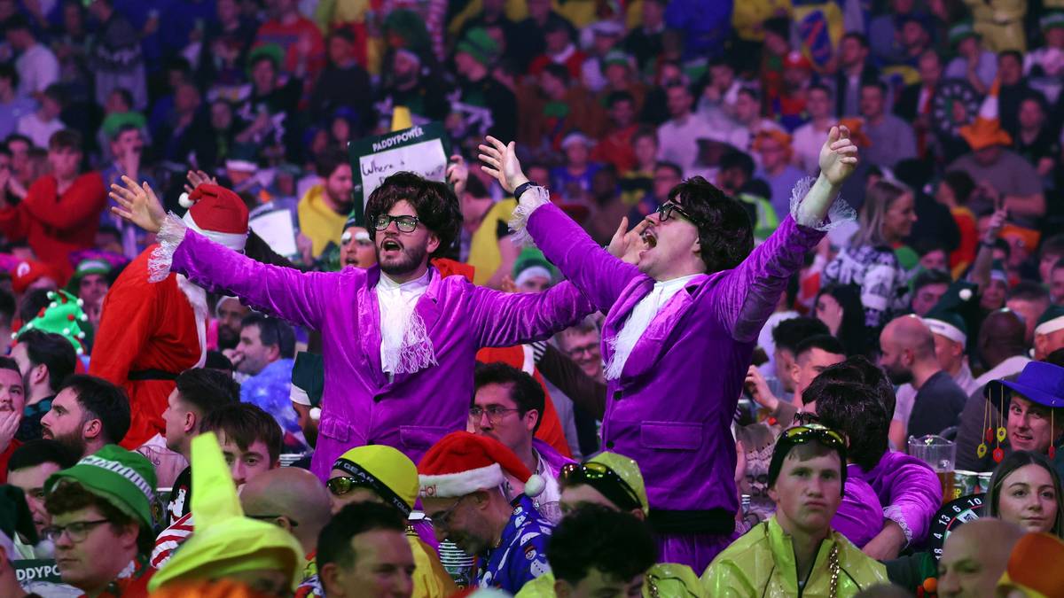 Widownia podczas zawodów darta, ubrana w kolorowe stroje i przebrania, z dwiema osobami w fioletowych marynarkach wznoszącymi ręce.