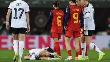 Piłkarki w białych i czerwonych strojach sportowych stoją wokół leżącej na murawie zawodniczki.