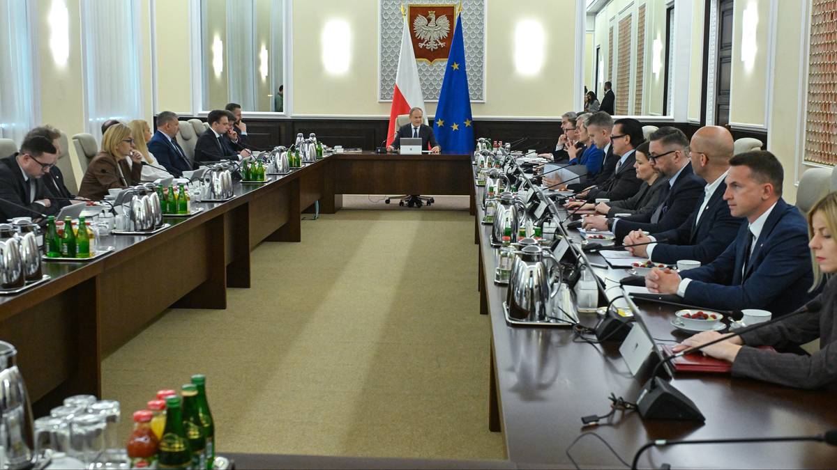 Grupa os&oacute;b siedzących przy długim stole w sali konferencyjnej, przed nimi ustawione są notatniki, długopisy i butelki z wodą. Na stole znajdują się r&oacute;wnież dzbanki z wodą.