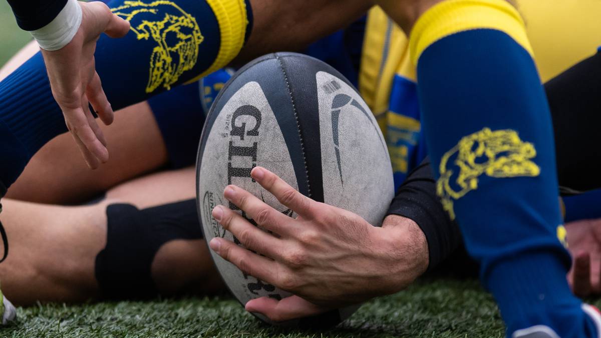 Piłka do rugby trzymana przez gracza, otoczona przez inne ręce i nogi zawodników w niebiesko-żółtych strojach.
