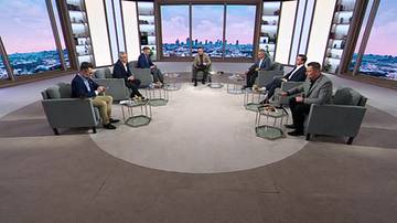 Ośmiu mężczyzn w garniturach siedzi w okręgu na sofach w studiu telewizyjnym z widokiem na miasto w tle.