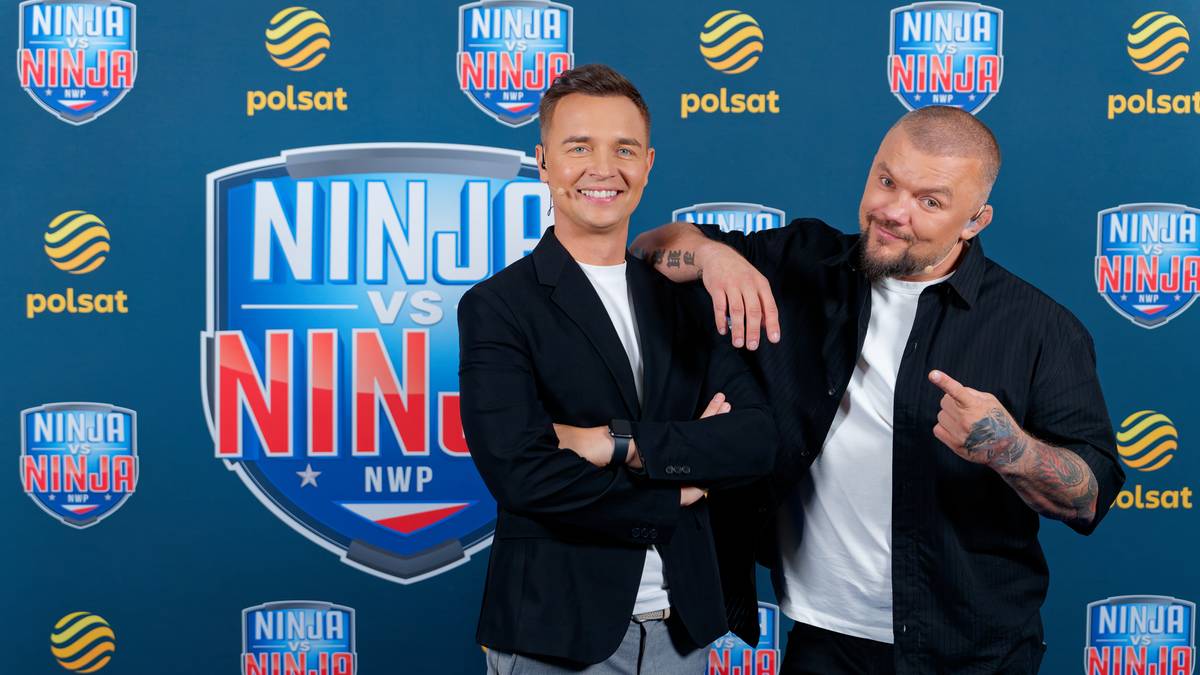 Dwóch mężczyzn stojących obok siebie. Po lewej stoi mężczyzna w czarnej marynarce i białej koszulce, uśmiechnięty. Po prawej stoi mężczyzna z tatuażami na ramionach, ubrany w czarną koszulę, z uniesionym palcem wskazującym.