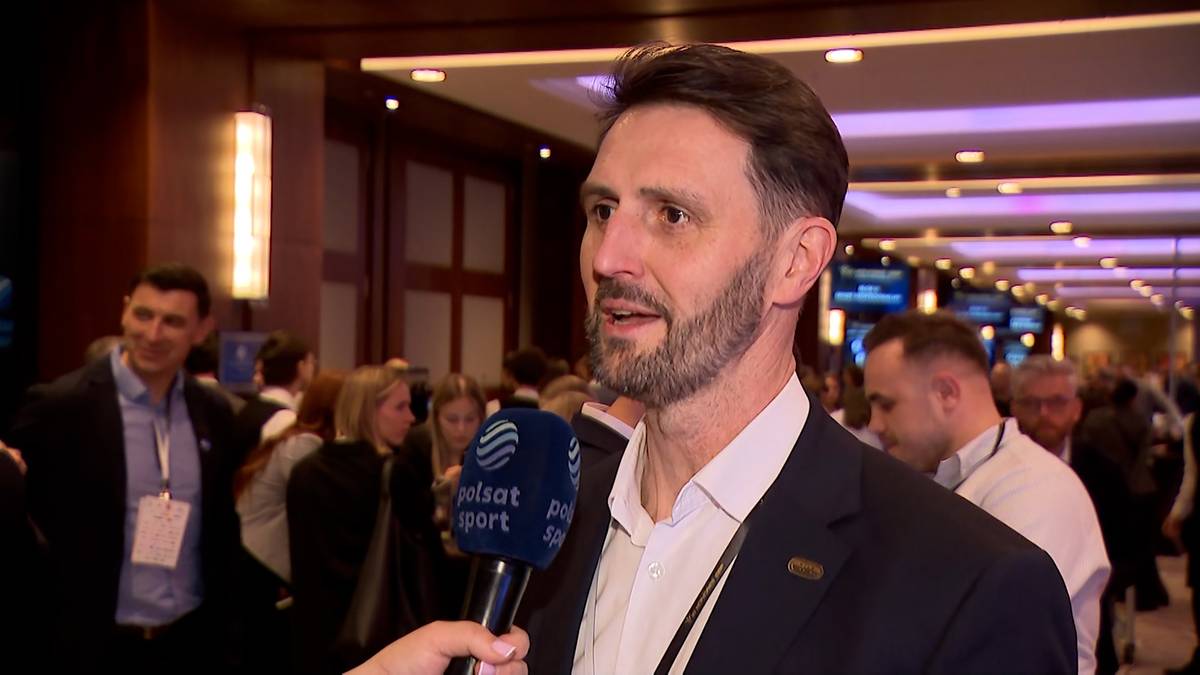 Mężczyzna z brodą i kr&oacute;tkimi włosami, ubrany w ciemną marynarkę i białą koszulę, udziela wywiadu w pomieszczeniu z innymi ludźmi w tle.