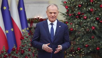 Donald Tusk w garniturze stoi przed choinkami i flagami Polski i Unii Europejskiej.