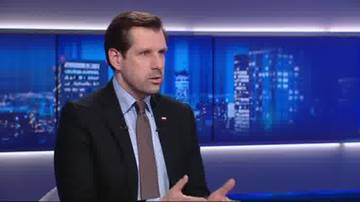 Mężczyzna w garniturze i krawacie m&oacute;wi, siedząc przed ekranami wyświetlającymi nocny widok miasta.