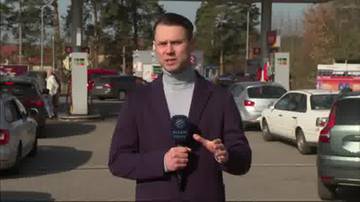 Mężczyzna dziennikarz z mikrofonem stoi na tle stacji paliw, na kt&oacute;rej widoczne są samochody i inni ludzie.