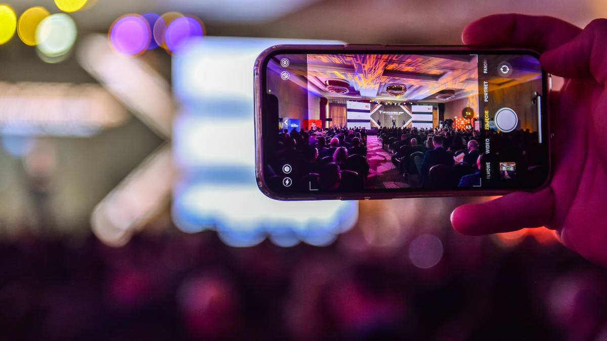 Dłoń trzyma smartfon nagrywający wnętrze sali konferencyjnej podczas wydarzenia z dużą liczbą uczestników i scenicznym oświetleniem.