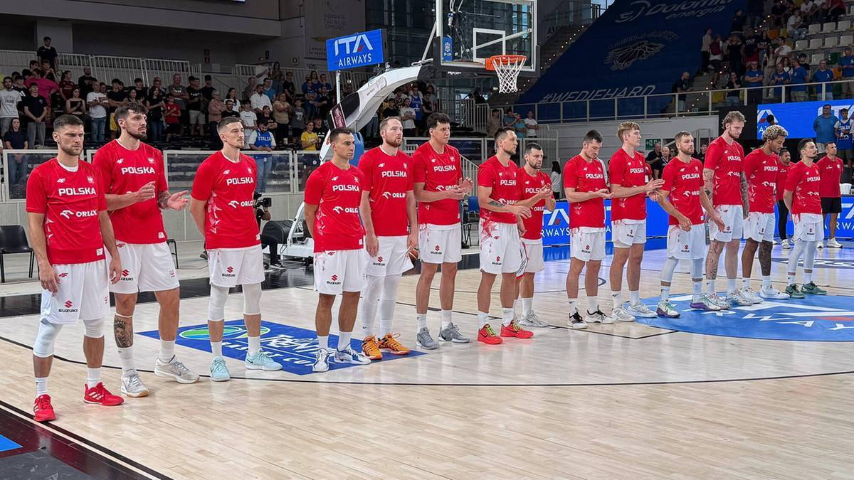 Drużyna polskich koszykarzy w czerwonych strojach stoi w rzędzie na boisku.