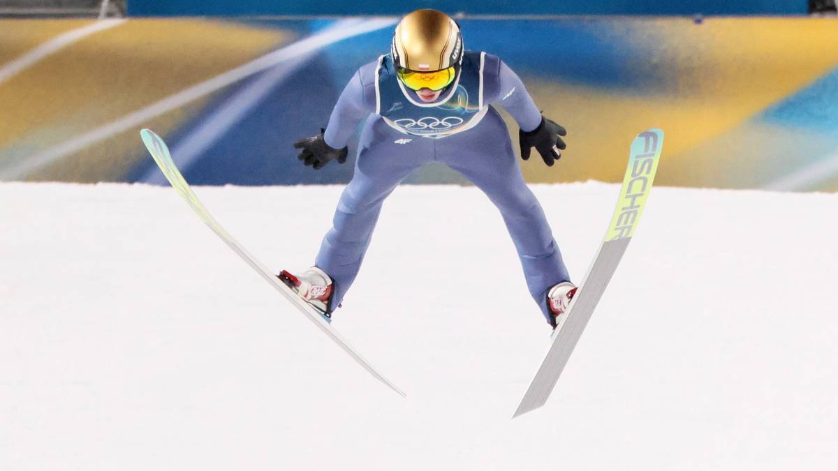 Skoczek narciarski w niebieskim kombinezonie i ż&oacute;łtym kasku w trakcie lotu na nartach, na tle niebieskiego baneru z pięcioma k&oacute;łkami symbolizującymi igrzyska olimpijskie.