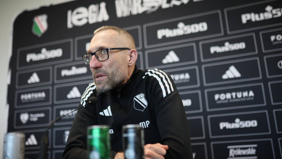 Trener Legii Warszawa podczas konferencji prasowej, siedzi za stołem z mikrofonem, w tle tablica sponsorska.