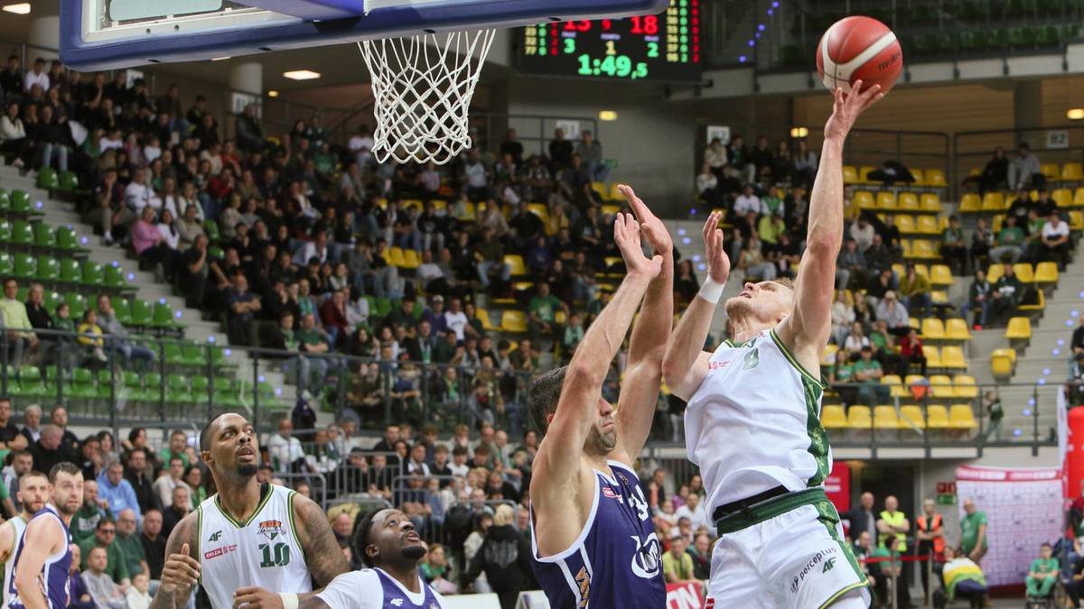 Koszykarz w białej koszulce z numerem 10 na plecach próbuje wykonać rzut do kosza, podczas gdy dwaj gracze drużyny przeciwnej próbują go zablokować. Na pierwszym planie widać zawodnika w białej koszulce i czarnych spodenkach. W tle widownia wypełniająca trybuny hali sportowej.