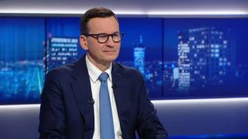 Mateusz Morawiecki w okularach, ubrany w granatowy garnitur i niebieski krawat, siedzi w studiu telewizyjnym.