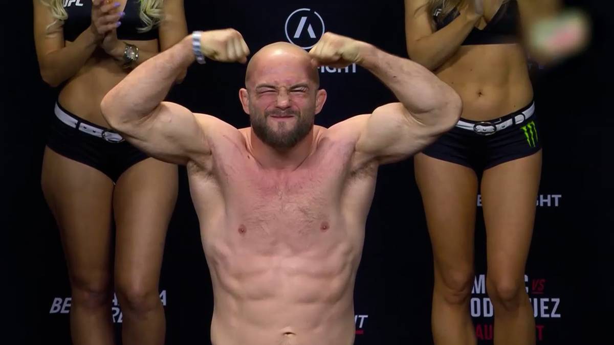 Zawodnik MMA z brodą, o napiętych mięśniach biceps&oacute;w, z zamkniętymi oczami i wykrzywioną twarzą, stoi na pierwszym planie. Po jego bokach, w połowie widoczne, stoją dwie kobiety w strojach sportowych.
