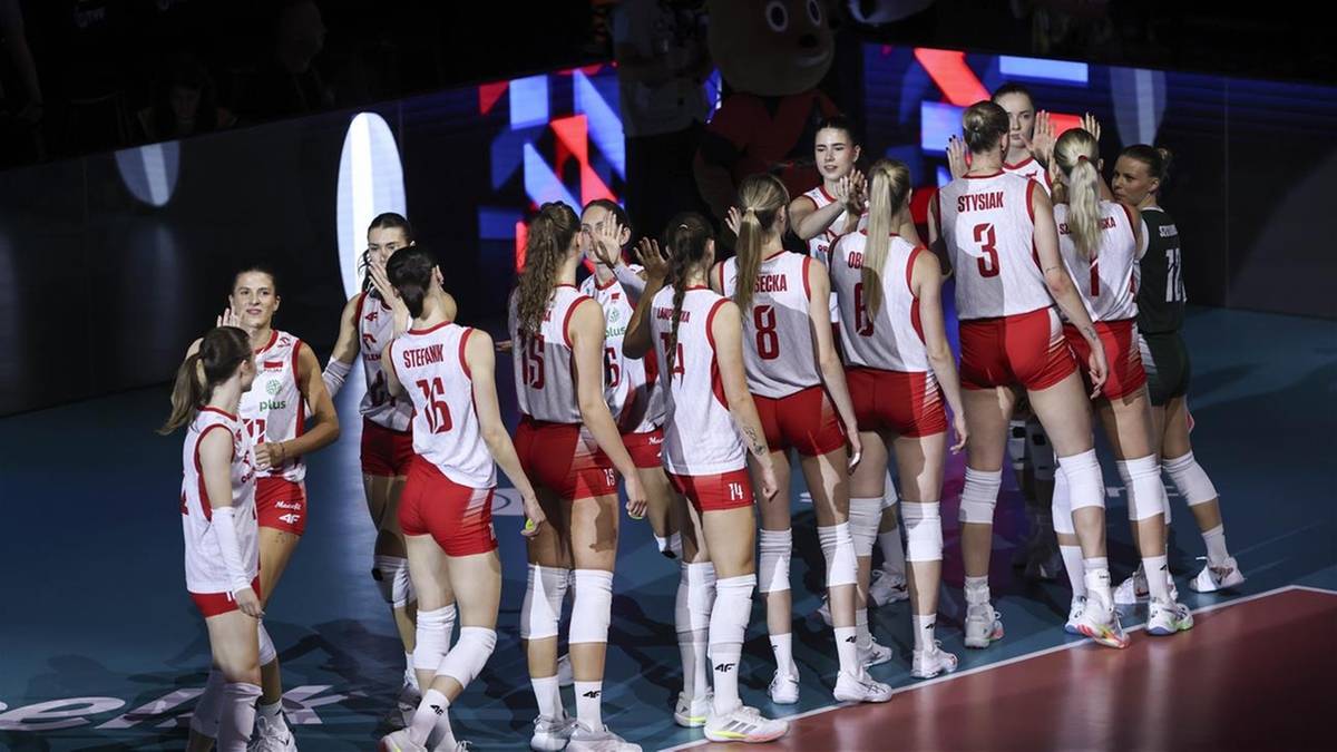 Drużyna siatkarek w białych i czerwonych strojach stoi na parkiecie hali sportowej.
