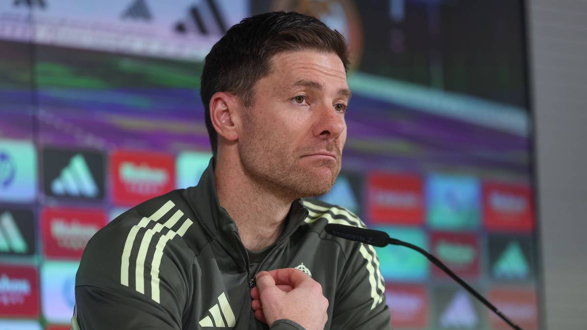 Xabi Alonso siedzi przy mikrofonie podczas konferencji prasowej, ubrany w szarą bluzę sportową z białymi paskami.