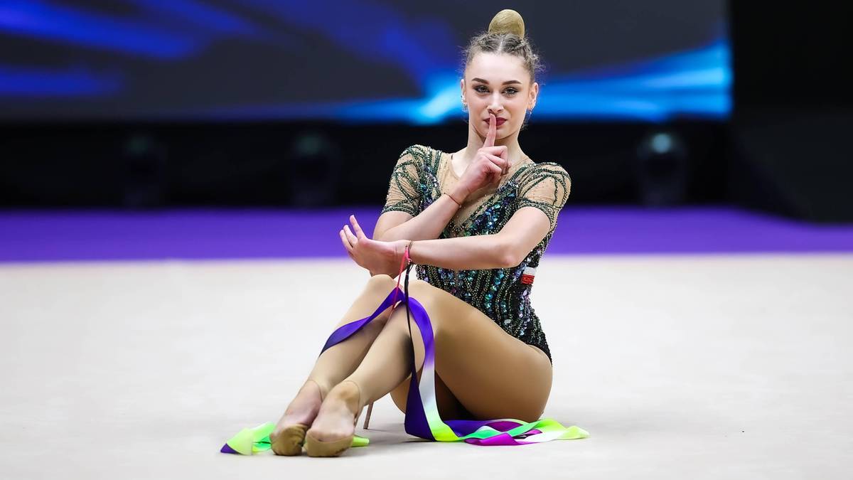 Młoda gimnastyczka rytmiczna siedzi na podłodze, trzymając w rękach kolorową wstążkę. Ma na sobie błyszczący, ciemny strój gimnastyczny.