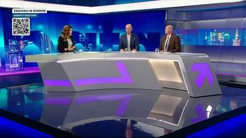 Trzy osoby siedzą za nowoczesnym stołem w studiu telewizyjnym, dyskutując. Na stole znajdują się mikrofony i materiały do pisania. W tle widać niebieskie i fioletowe oświetlenie.