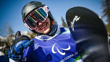 Zawodniczka snowboardu z uśmiechem na twarzy pokazuje kciuk w g&oacute;rę, trzymając deskę snowboardową.