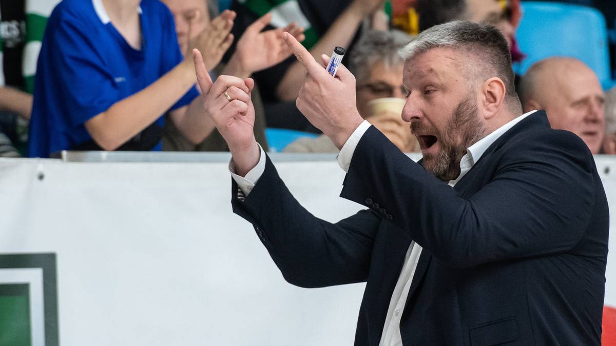 Trener drużyny koszykarskiej w garniturze i białej koszuli, z uniesionymi palcami wskazującymi, krzyczy z ekscytacją, stojąc przed publicznością.