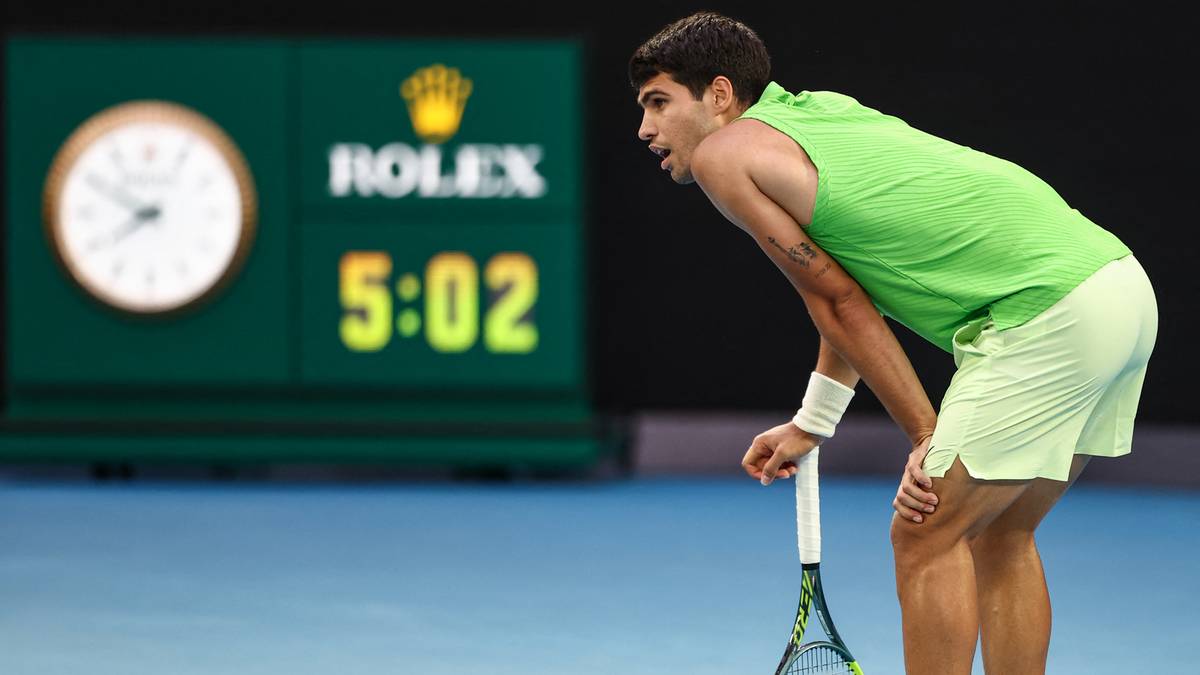Młody mężczyzna w zielonej koszulce i spodenkach tenisowych pochyla się z rakietą tenisową, patrząc na zegar z napisem ROLEX i wynikiem 5:02.