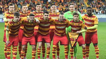 Drużyna piłkarska Jagiellonii Białystok w komplecie, stojąca na murawie stadionu, ubrana w czerwono-ż&oacute;łte stroje. Jeden z zawodnik&oacute;w trzyma proporzec.