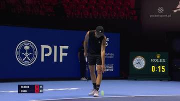 Tenisista Alexander Blockx podczas meczu, pochylony, aby podnieść piłkę tenisową. W tle widać tablicę wynik&oacute;w.