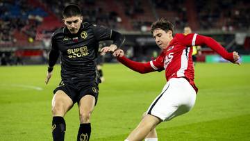 Dwóch piłkarzy w akcji na boisku, jeden w czarnej koszulce z żółtymi elementami, drugi w czerwonej koszulce z białymi spodenkami.