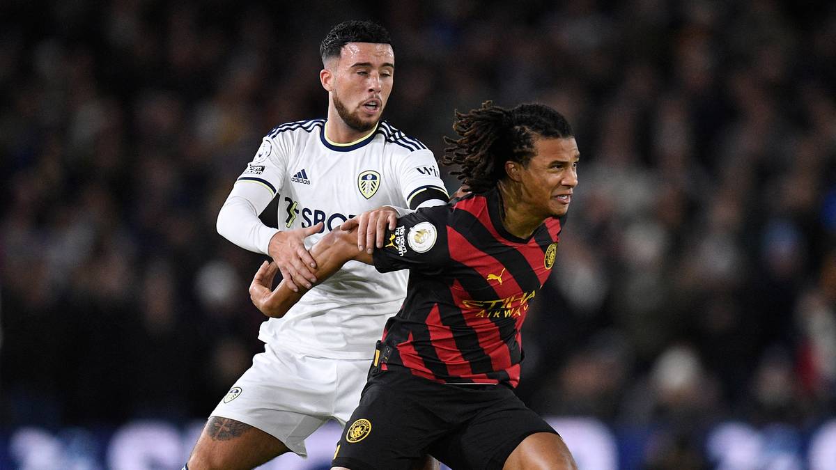 Dwóch piłkarzy na boisku podczas meczu, jeden w białej koszulce Leeds United, drugi w czarno-czerwonej koszulce.