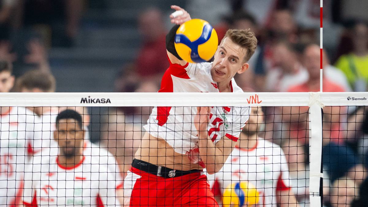 Siatkarz w białej koszulce z czerwonymi elementami i spodenkach, wykonuje zagrywkę piłką siatkową w żółto-niebieskich barwach. Na pierwszym planie siatka z logo Mikasa.