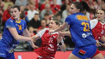 Trzy zawodniczki piłki ręcznej podczas meczu, dwie w niebieskich strojach atakują zawodniczkę w czerwonym stroju.