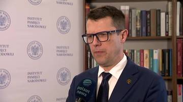 Mężczyzna w okularach i garniturze, stojący przed ścianą z logo Instytutu Pamięci Narodowej. Trzyma mikrofon Polsat News.