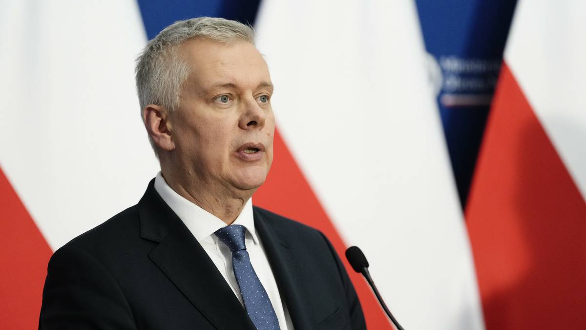 Mężczyzna w garniturze i krawacie mówi do mikrofonu na tle flag Polski.