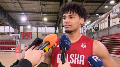 Młody mężczyzna w czerwonej koszulce z napisem "POLSKA" otoczony przez mikrofony, udzielający wywiadu w hali sportowej.