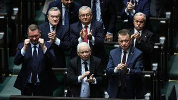 Grupa posł&oacute;w w garniturach siedząca w ławach sejmowych, klaszcząca.