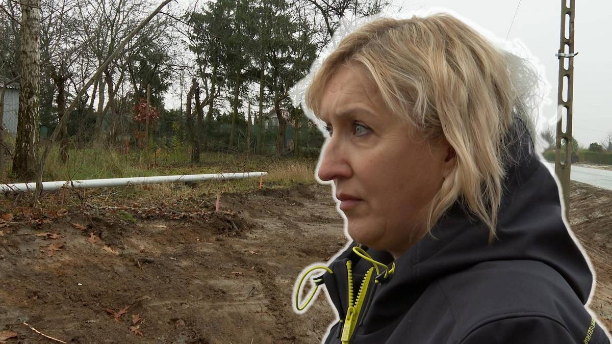 Kobieta o blond włosach patrzy w bok z zamyślonym wyrazem twarzy, na tle wyciętych drzew i terenu budowy.