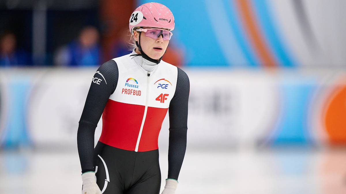 Łyżwiarka szybka w różowym kasku i okularach, w biało-czerwonym stroju sportowym, stoi na lodowisku.