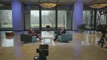 Dwie osoby siedzą naprzeciwko siebie na kanapach w przestronnym, nowoczesnym studiu telewizyjnym. Przed nimi stoi niski stolik.