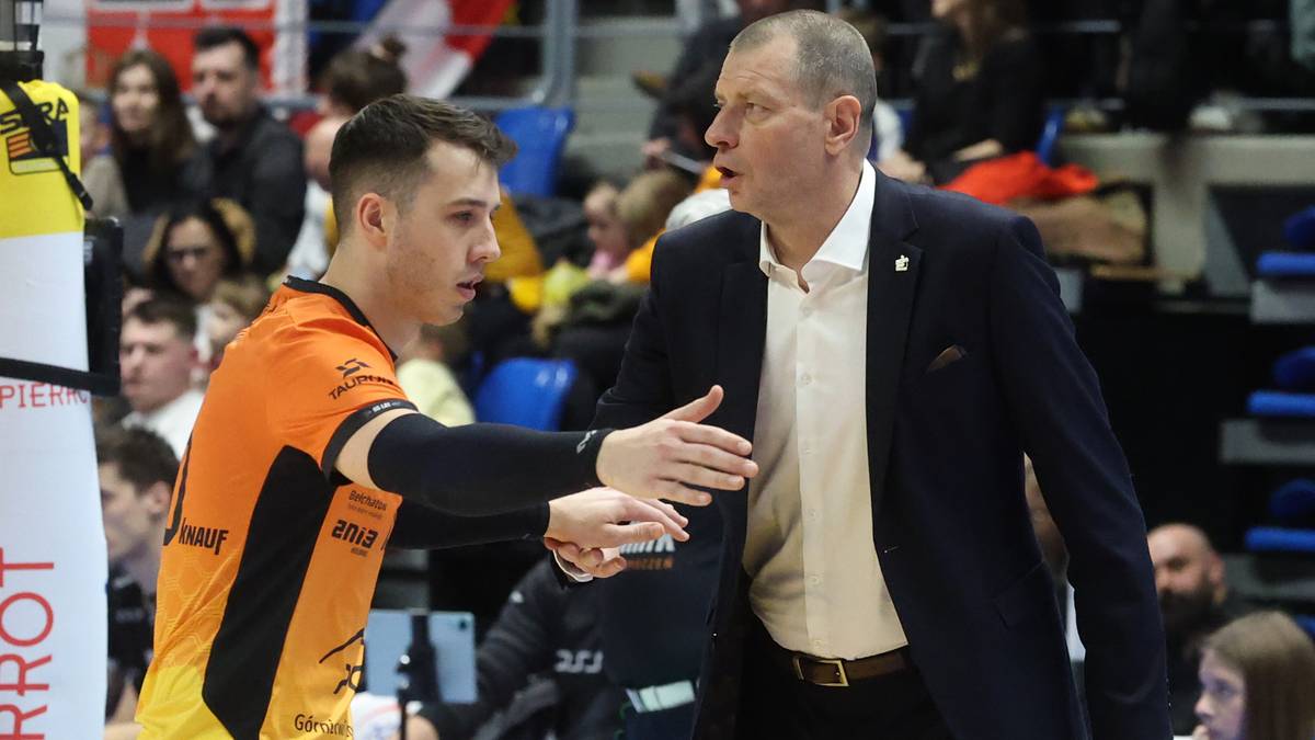 Dw&oacute;ch mężczyzn, jeden w pomarańczowo-czarnej koszulce sportowej, drugi w granatowym garniturze i białej koszuli, rozmawia podczas meczu siatk&oacute;wki.