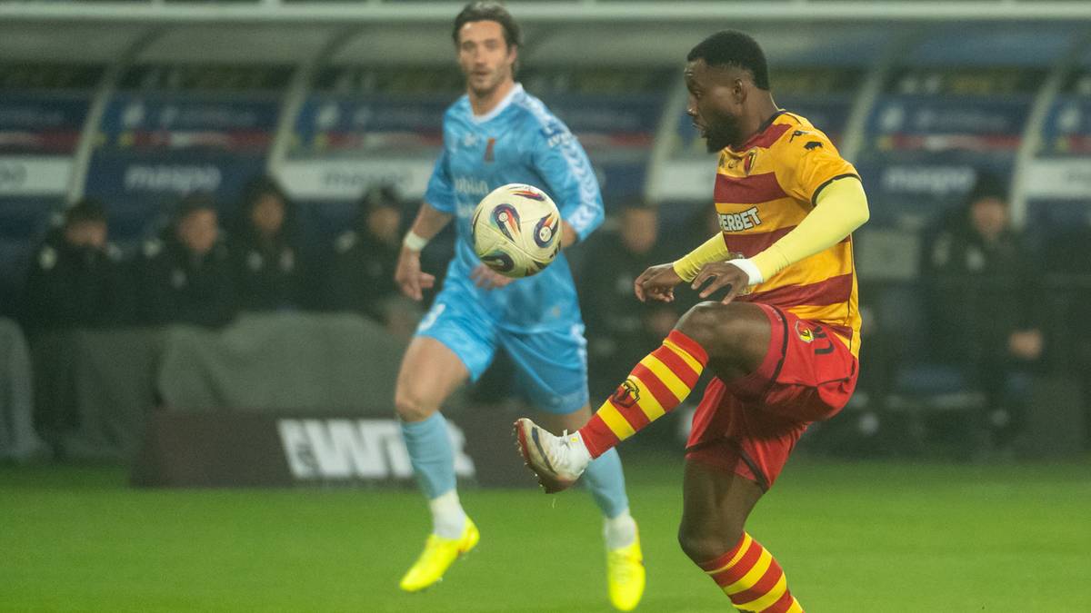 Dwóch piłkarzy na boisku, jeden w czerwono-żółtej koszulce próbuje odebrać piłkę zawodnikowi w niebieskim stroju.