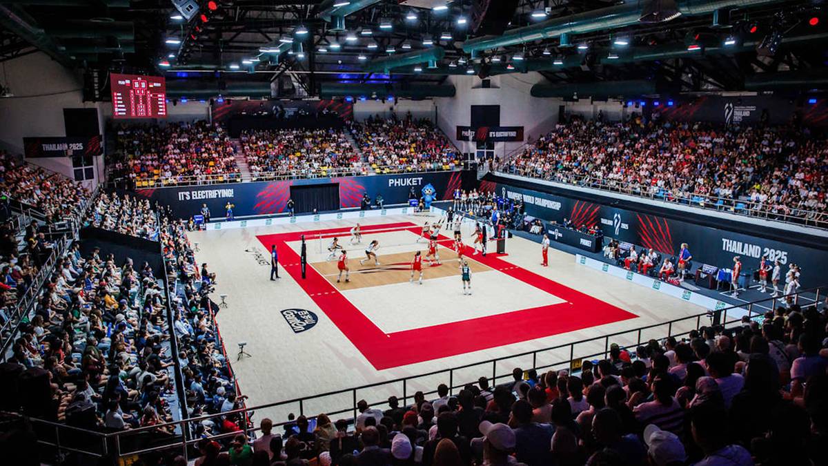 Siatkarki w czerwonych strojach grają w siatkówkę na boisku z czerwonym obrzeżem, otoczone przez trybuny wypełnione widzami w dużej, jasnej hali sportowej.